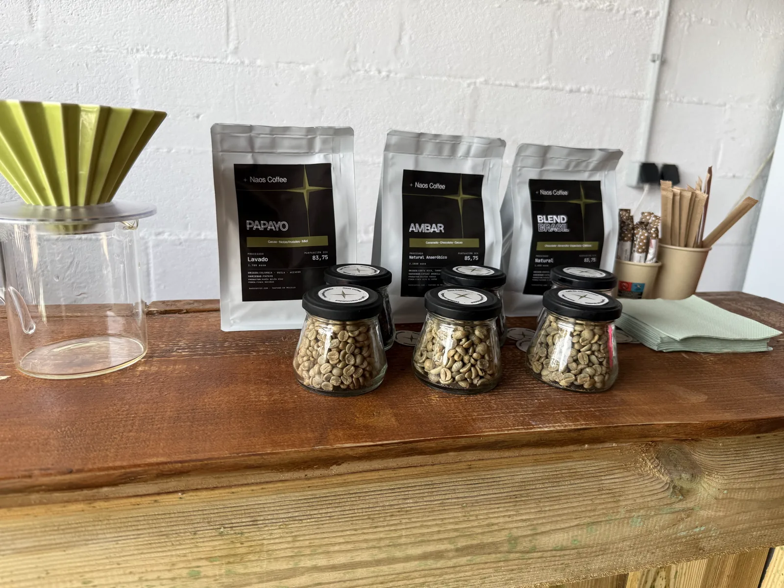 Bolsas de los cafés Papayo, Ámbar y Blend de Naos Coffee junto a tres tarros de café verde y un dripper V60 sobre la barra.
