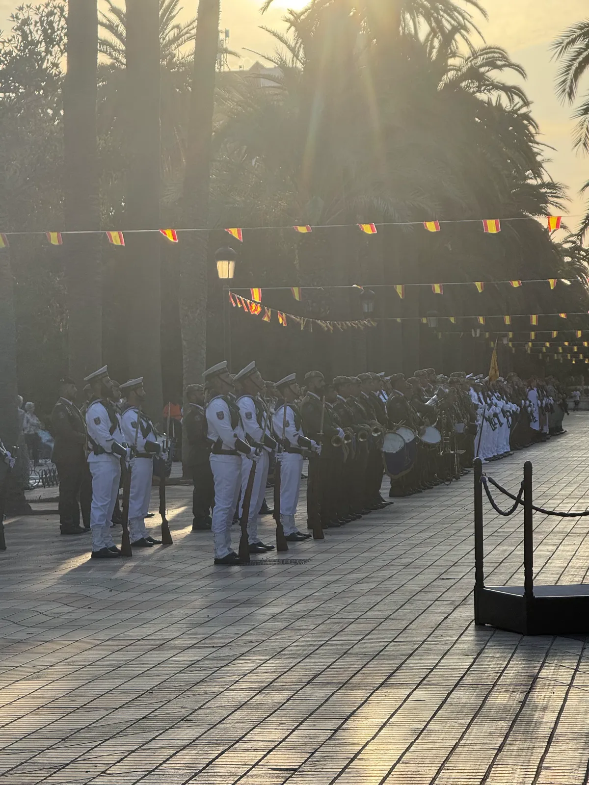 Banda de música uniformada en el paseo central del Parque Hernández de Melilla al caer la tarde, con banderines sobre las palmeras.