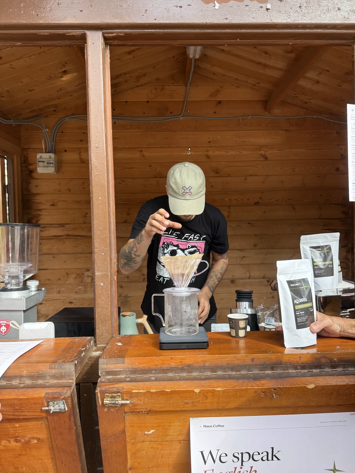 Barista de Naos Coffee preparando un café de filtro en V60 dentro de la caseta, con un cartel de «We speak English» en primer plano.