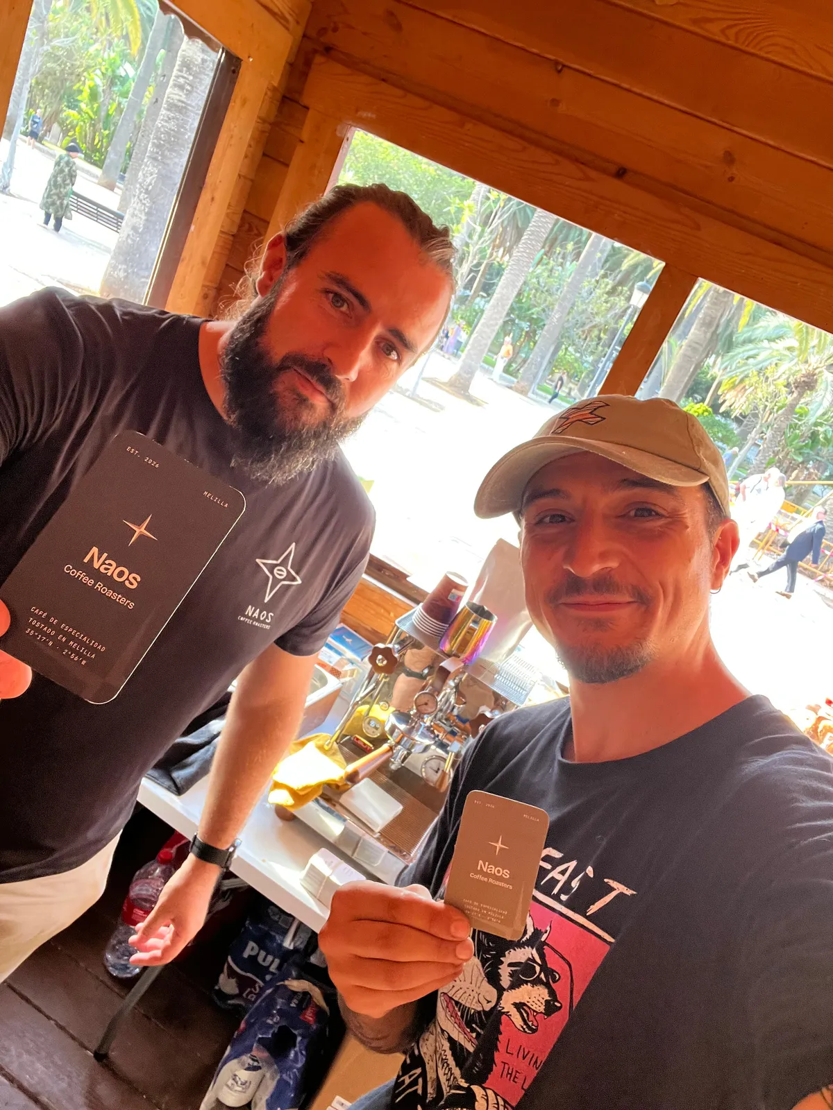 Dos personas sonríen dentro de la caseta de Naos Coffee enseñando las tarjetas de la marca.