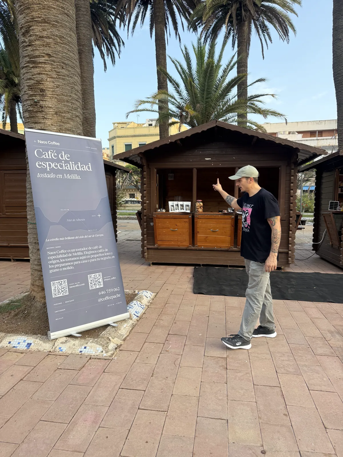Barista de Naos Coffee señalando la caseta recién montada en el Parque Hernández, con el roll-up de la marca bajo las palmeras.